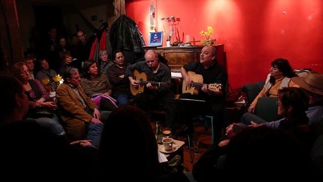 wunderschönes Photo im Café Esperanza. Kai Heumann und Sergio Mansilla mit ihren Gitarren vor dem Caféklavier an der roten Wand. Das Café ist proppenvoll und die Leute sitzen (im Hintergrund stehen) im Hufeisen um die Musiker herum